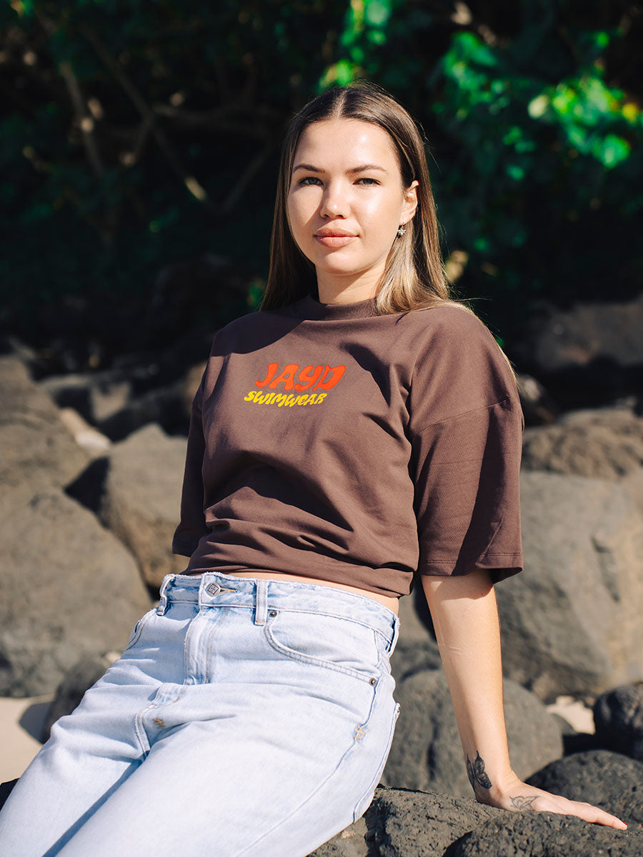 Front detail of JAYD Swimwear's brown oversized tee, printed with a retro-style orange JAYD and yellow Swimwear logo.
