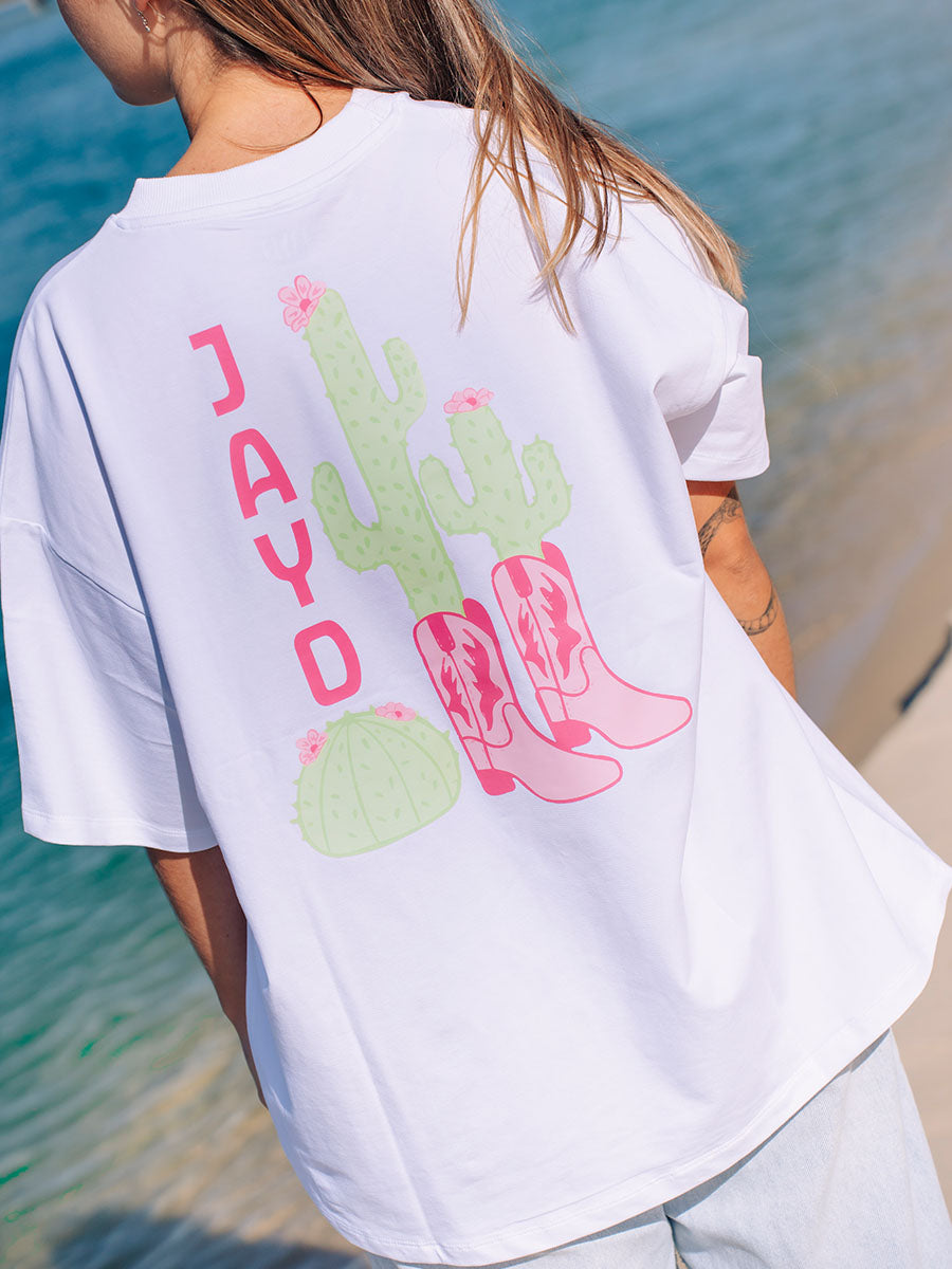 Woman wearing a white JAYD Swimwear oversized boxy tee with a pink cowgirl boots and cactus graphic print on the back, styled at the beach.