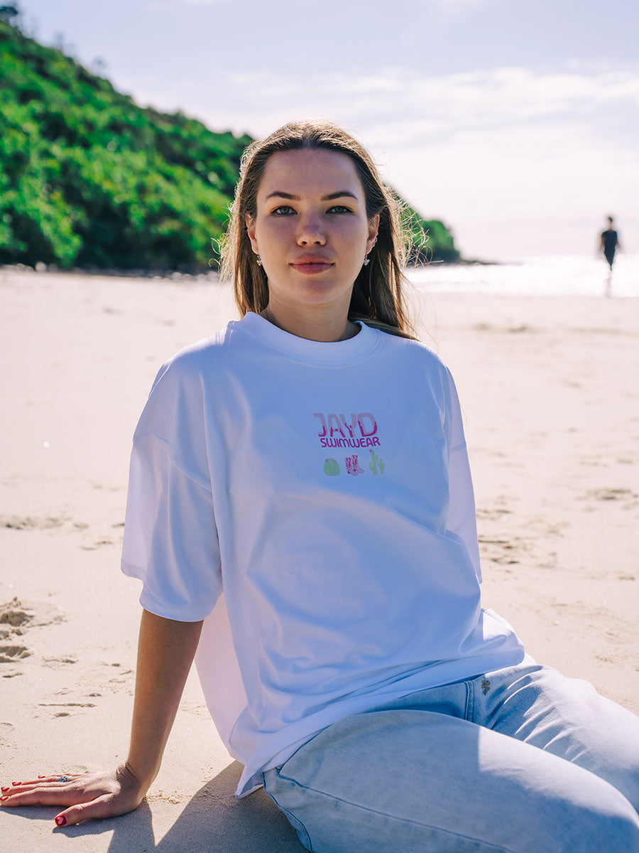 White oversized JAYD Swimwear t-shirt with a pink cowgirl boots and cactus back print, a fun boxy fit tee for beach days and lounging.