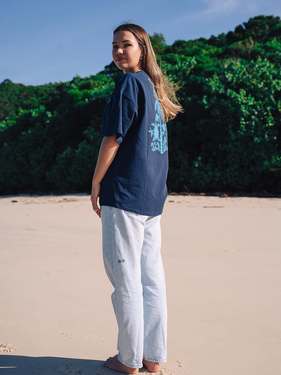 Woman wearing a navy JAYD Swimwear oversized boxy tee with a light blue tropical graphic print on the back, paired with light wash denim jeans on the beach.