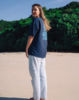 Woman wearing a navy JAYD Swimwear oversized boxy tee with a light blue tropical graphic print on the back, paired with light wash denim jeans on the beach.