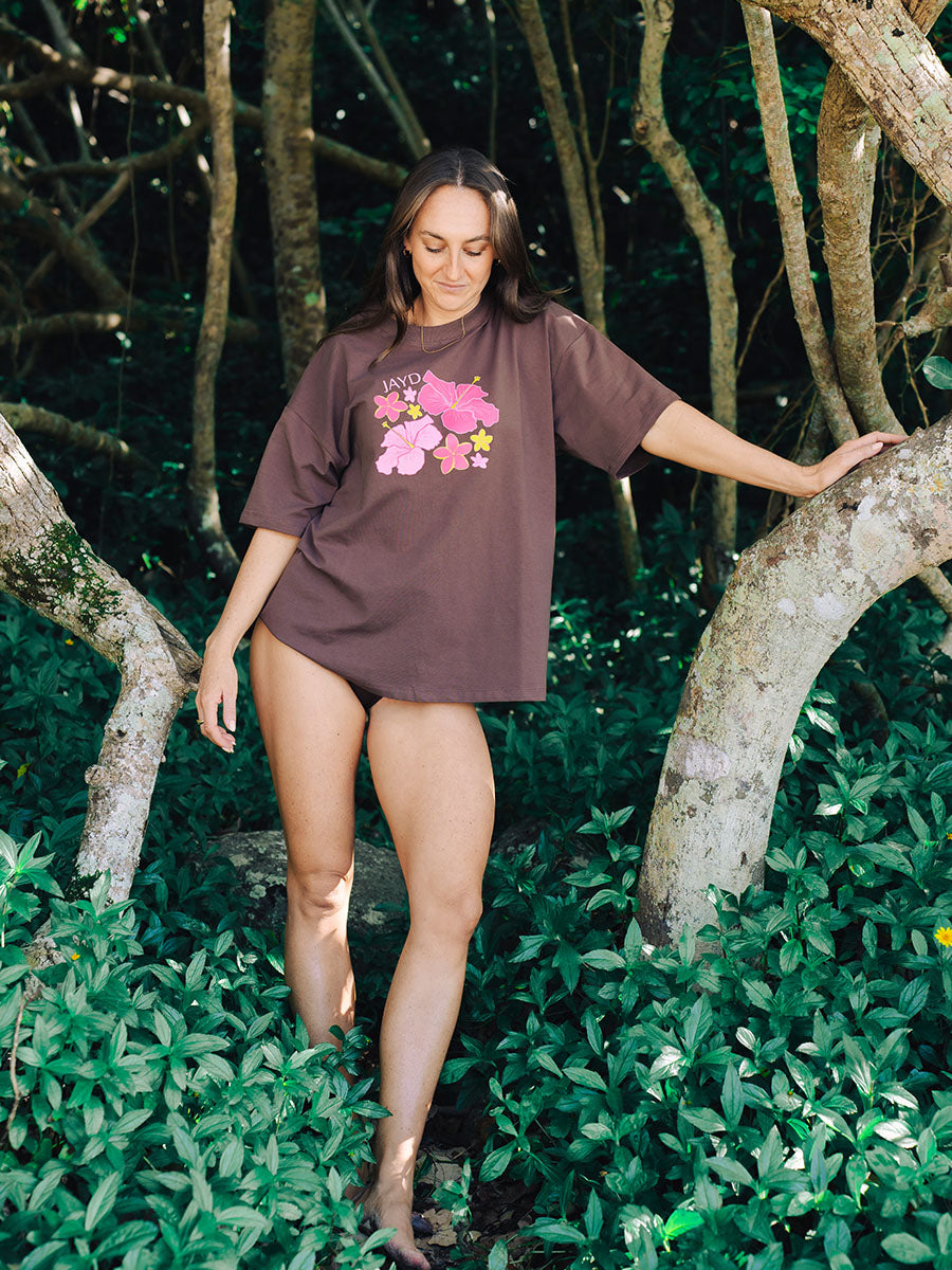 Woman wearing a brown JAYD Swimwear oversized boxy tee with a pink and yellow hibiscus floral graphic print on the front, styled in a tropical forest setting.