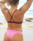 Back strap view of a female swimmer in a JAYD Swimwear Torquay bikini top in Sangria and pink bikini bottoms, standing on a beach.