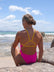 Back view of surf athlete, Milly Derbyshire, sitting on a beach wearing a JAYD Swimwear Zambia One Piece in Pink Sorbet with yellow straps.