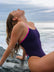 Side view of model on a rock wearing JAYD Swimwear athletic one piece, Lennox, in Acai Purple.