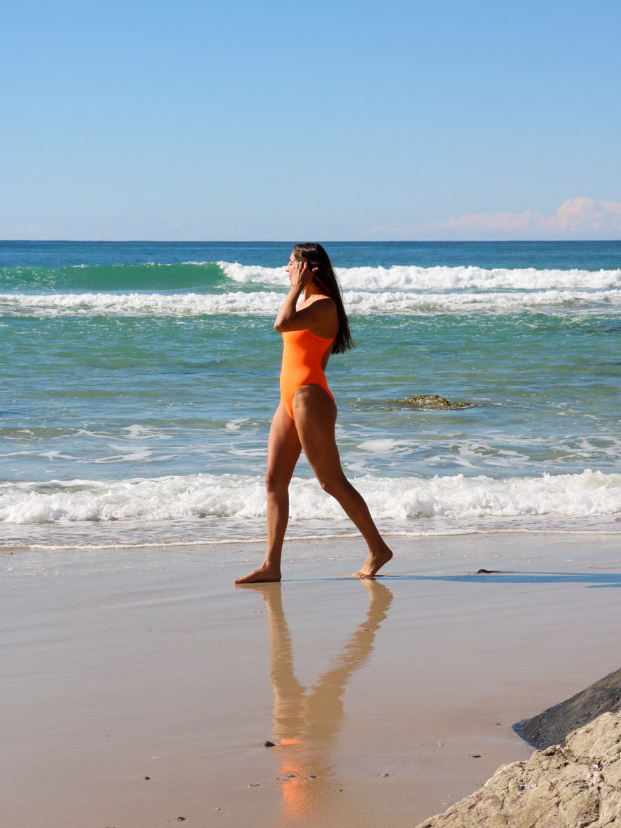 Side view of model walking on a beach wearing JAYD Swimwear athletic one piece Apsley in Neon Orange.