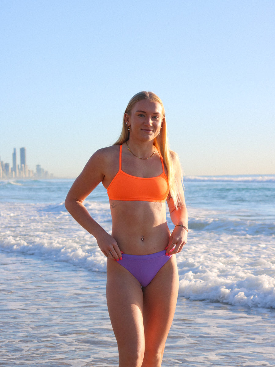 Front view of model on a beach wearing JAYD Swimwear athletic bikini top Torquay in Neon Orange.