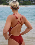 Close back strap view of a female athlete wearing a JAYD Swimwear Finley swimsuit in Cinnamon, standing on a beach with ocean in the background.