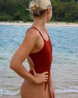 Side view of a female athlete wearing a JAYD Swimwear Finley swimsuit in Cinnamon, standing on a beach with ocean in the background.