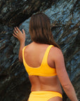 Back view of Australian triathlete, Charlotte Derbyshire, wearing a Sunshine yellow JAYD Swimwear bikini top against a rocky background