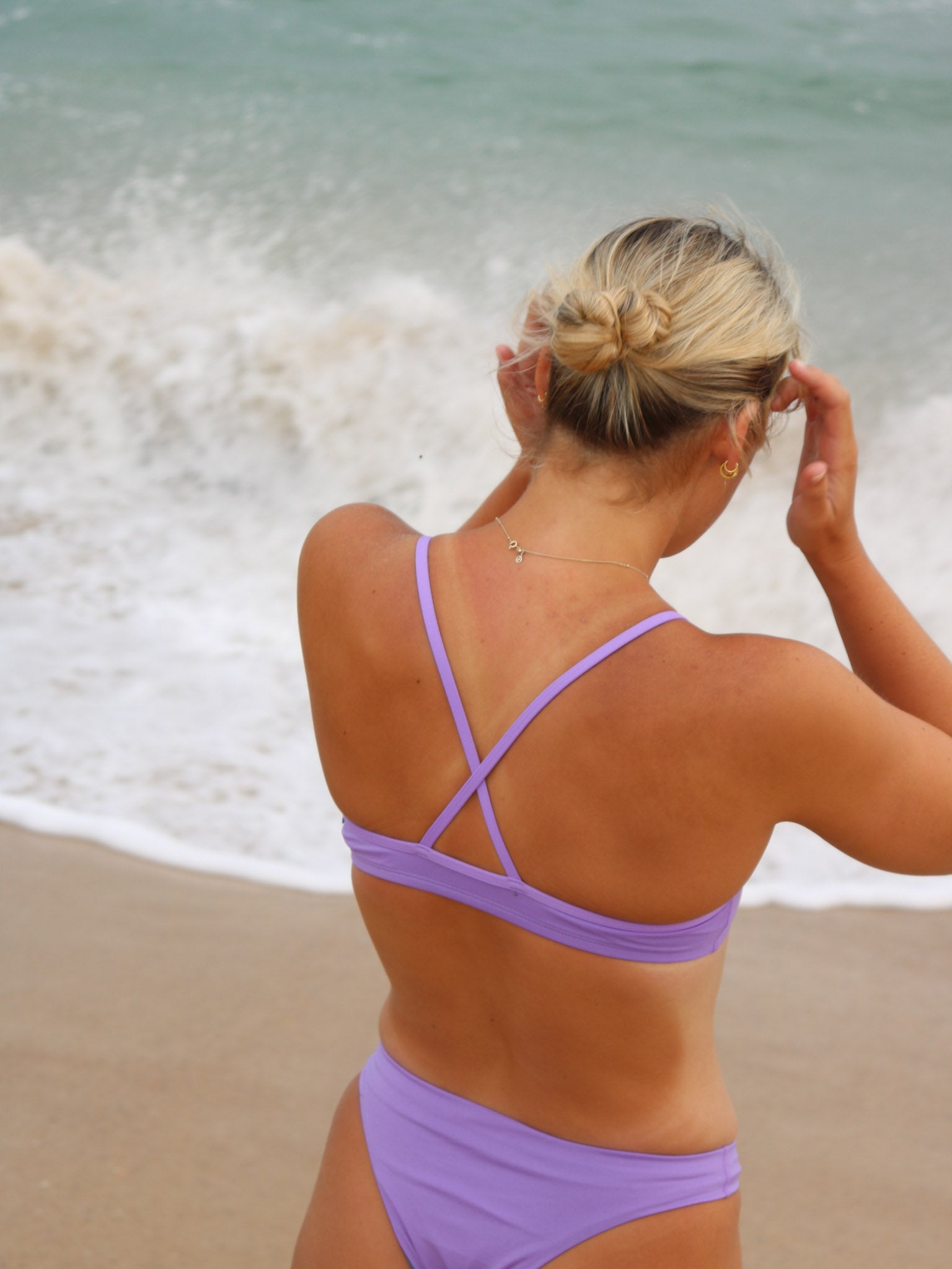 Close back strap view of a female athlete in a JAYD Swimwear Torquay bikini top in lilac haze, standing on a beach with waves in the background.