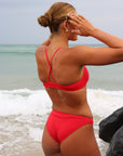 Back view of a female athlete in a JAYD Swimwear Torquay bikini top in Ruby, standing in front of waves on the beach.