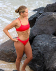 Front view of a female athlete in a JAYD Swimwear Torquay bikini top in Ruby, standing at the beach.