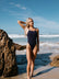 Athlete in a Acai JAYD swimsuit standing on a rocky beach with ocean waves in the background