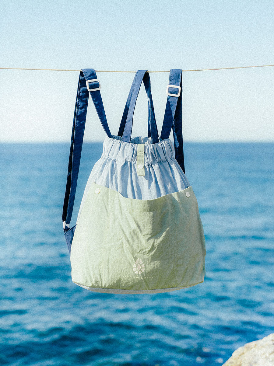 A light blue and pale green drawstring backpack with dark blue shoulder straps hanging from a wire over the ocean.