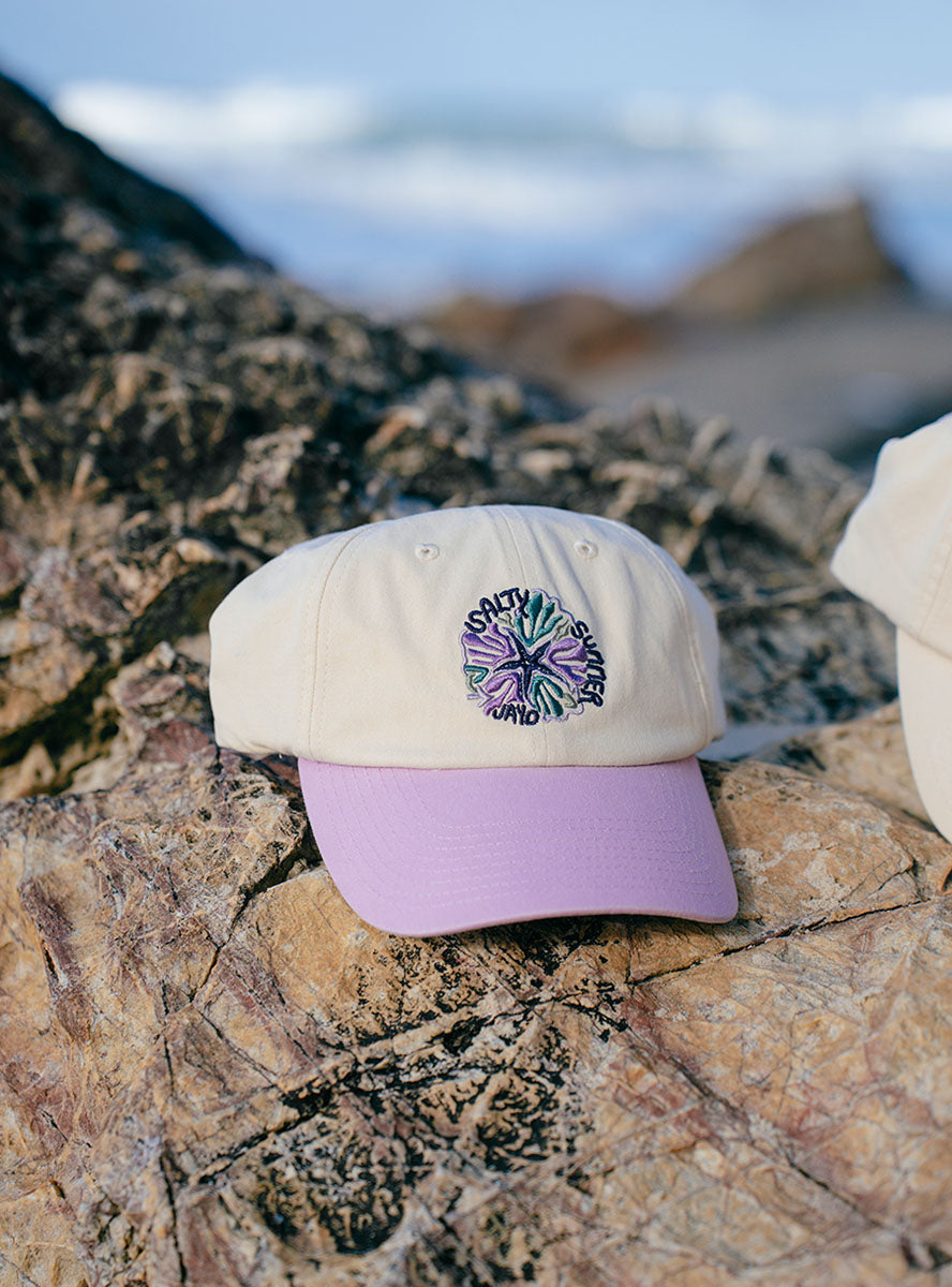 Cream and purple JAYD Swimwear Baseball Cap sitting on a rock at the beach.