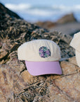 Cream and purple JAYD Swimwear Baseball Cap sitting on a rock at the beach.