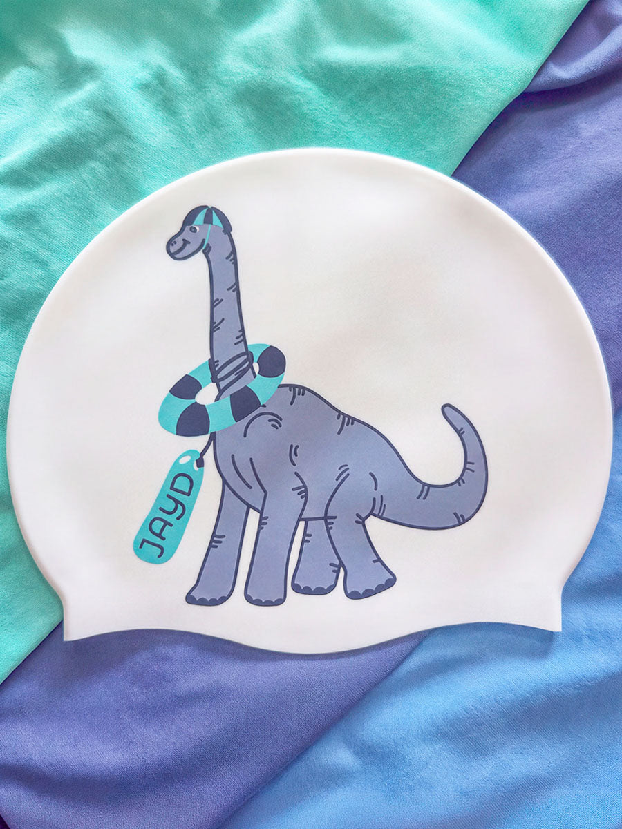 A white swim cap featuring a cartoon Brachycephalus dinosaur in gray with a teal and black striped swim ring and a teal tag that reads 'JAYD', laid flat on a draped background of teal and blue fabric.
