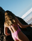 Person in a fairy floss pink one piece standing on rocks by the ocean. She is wearing a chlorine resistant one piece built for performance.