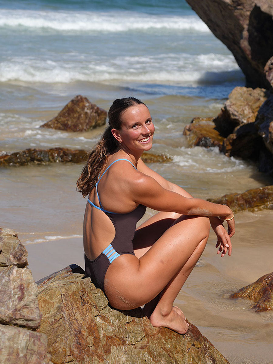 Molly enjoying a day at the beach basking in the sunshine on a rock with the ocean in the background, wearing the JAYD Lennox One Piece swimsuit.