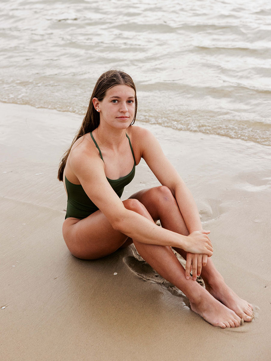JAYD Swimwear model Cyra wearing the tie-back Olive One Piece in Riva, sitting on the sand relaxing at the beach.