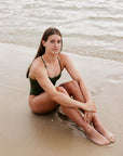 JAYD Swimwear model Cyra wearing the tie-back Olive One Piece in Riva, sitting on the sand relaxing at the beach.