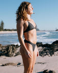 Woman in a Carbon grey bikini standing on a beach with ocean and sky in the background. She wears the Triangle top and Cali bottom