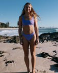 Athlete Brodee in JAYD Swimwear's Lilac haze bikini standing on a beach with ocean and skyline in the background. Brodee wears the Tahiti bottom and Nalu top.
