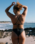 Person wearing a Carbon grey mid coverage bikini bottom, called Cali, standing on a beach with ocean and sky in the background. 