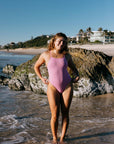Morgan in the TALLOW One Piece swimsuit in Fairy Floss pink, standing on a beach with rocks and water.