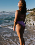 Woman in JAYD Swimwear's TALLOW One Piece in the shade Fairy Floss (light pink), standing on a beach with ocean waves in the background