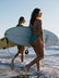 Two women walking on a beach with surfboards, one wearing the TALLOW One Piece swimsuit in Atlantic green.