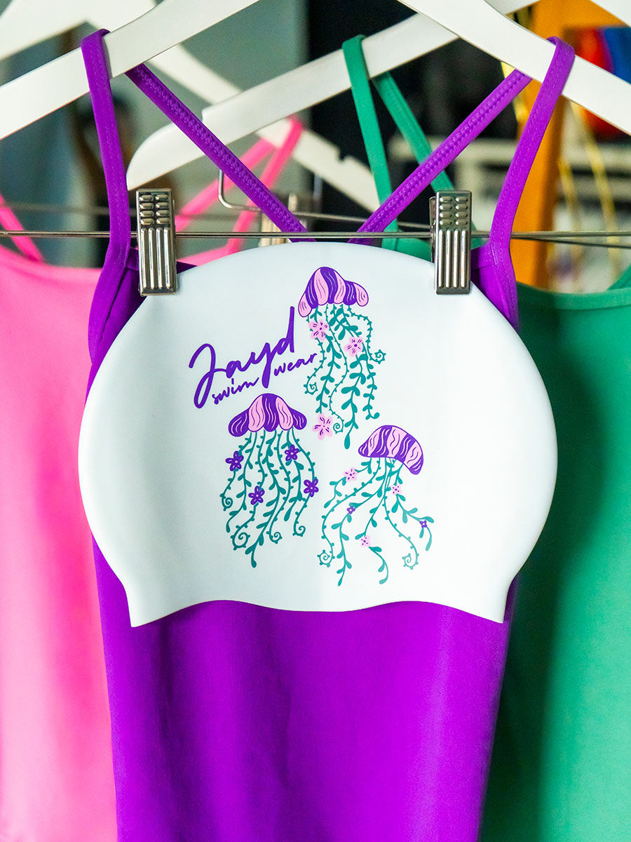 Three jellyfish flower illustrations on a white swim cap with pink, purple and green swimsuits in the background.