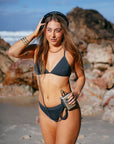 JAYD model Cass walking on a beach listening to a vintage music player, wearing the Kiama bikini top in carbon with matching carbon bottoms