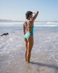 JAYD Swimwear model Sophie wearing the nordic one piece swimsuit with contrasting navy straps, standing in ankle deep water on the beach with waves breaking in the distance.