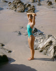 Side view of model Sophie wearing the JAYD Swimwear Lennox one piece in Nordic, standing on a rocky beach looking off into the distance.
