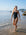 JAYD Swimwear model Emily walking in shallow ocean water on a beach wearing the Lennox one piece swimsuit in black with contrasting orange shoulder straps.