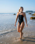JAYD Swimwear model Emily walking in shallow ocean water on a beach wearing the Lennox one piece swimsuit in black with contrasting orange shoulder straps.