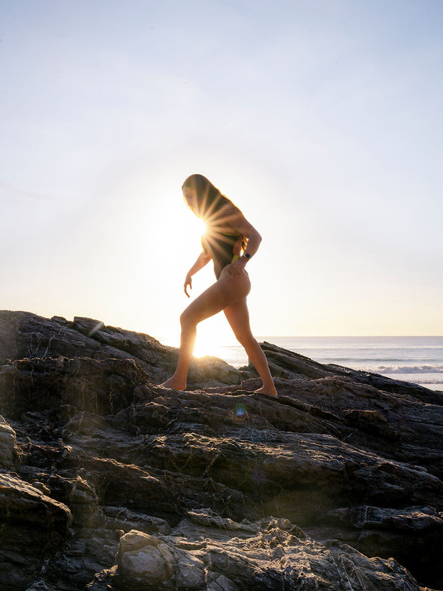 JAYD Swimwear model in the Lennox one piece swimsuit walks barefoot across dark rocky shoreline at sunset, with the sun low on the horizon and ocean in the background.