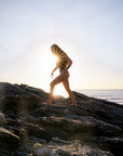 JAYD Swimwear model in the Lennox one piece swimsuit walks barefoot across dark rocky shoreline at sunset, with the sun low on the horizon and ocean in the background.