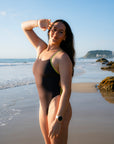 JAYD model Jenaya wearing the one-piece Lennox swimsuit in cacao brown with contrasting kiwi green shoulder straps, stands on a sandy beach with the ocean and rocks in the background.