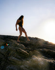 JAYD Swimwear modelwearing the Lennox one-piece swimsuit walks barefoot along a rocky shoreline at sunset, with the sun low on the horizon and lens flare visible.