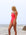 JAYD Swimwear model stands on a beach wearing a bright fiesta one-piece Lennox swimsuit with Atlantic shoulder straps. The background shows a calm sea and a pale, overcast sky.