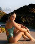 JAYD model Emily sitting on a beach at sunrise with seaweed covered rocks and the cityscape in the distance. She wears the Lennox one piece swimsuit in matcha green with contrasting storm blue shoulder straps and hip cut-outs.