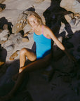 Model Emma sitting on rocky sand at a beach wearing the JAYD Lennox one piece swimsuit in nordic blue with orchid pink shoulder straps.
