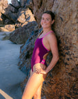 JAYD Swimwear athlete Charlotte Derbyshire leaning against a rock wearing the Lennox one piece swimsuit in sangria with contrasting orchid shoulder straps and 5 hip panel cut-outs.
