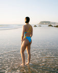 JAYD Swimwear model stands in shallow ocean water on a beach, viewed from behind, wearing the Lennox one-piece swimsuit with a bright blue Waikiki base and yellow Daffodil straps and side accents. The background shows calm water, distant rocks, and a pale sky.