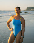Ski athlete Jenaya Massie standing on a beach at sunrise wearing the JAYD Lennox one piece in Waikiki blue with contrasting Daffodil yellow shoulder straps.