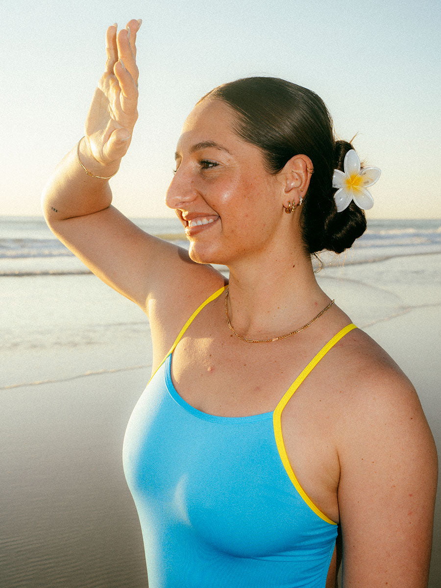 JAYD Swimwear model Jenaya standing on a beach wearing a bright blue Waikiki Lennoxone-piece swimsuit with yellow trim. She has a white frangipani flower clip in her hair, and the ocean and sand are visible in the background.