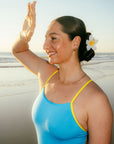 JAYD Swimwear model Jenaya standing on a beach wearing a bright blue Waikiki Lennoxone-piece swimsuit with yellow trim. She has a white frangipani flower clip in her hair, and the ocean and sand are visible in the background.